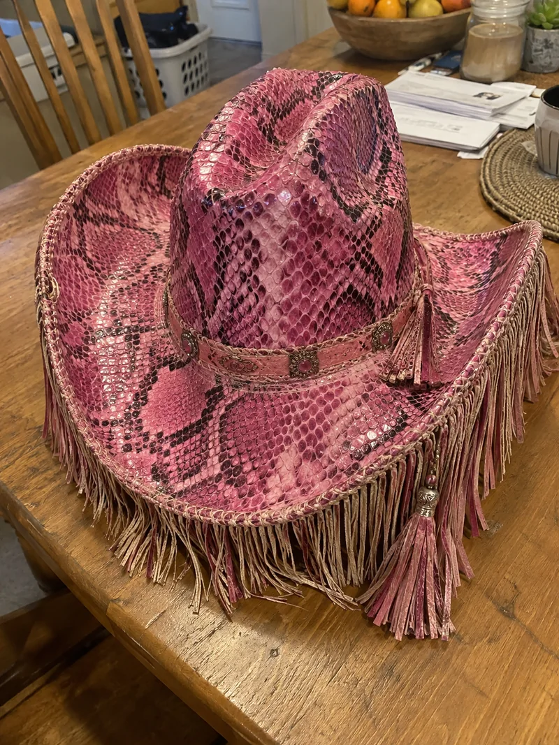 Pink cowboyhat i slangeskindsmønster med læderfrynser og kvaster, på et træbord.