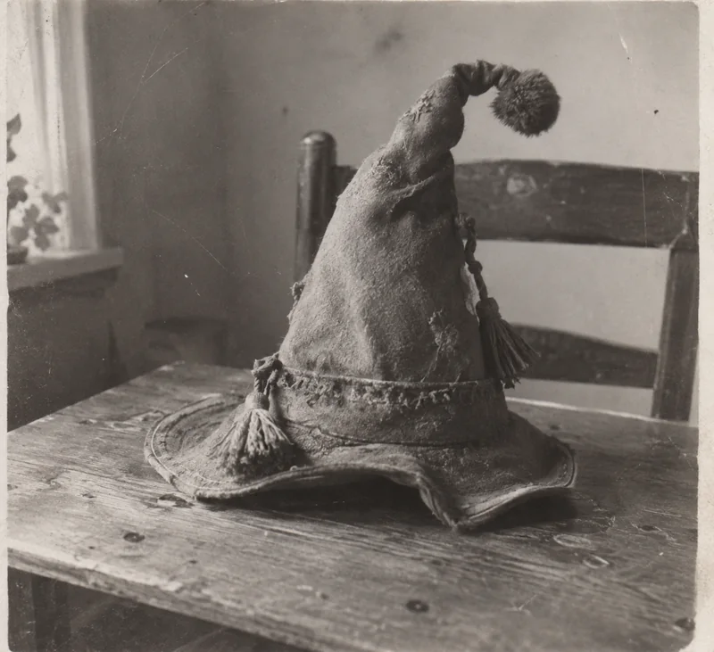 Gammelt sort-hvidt fotografi af en slidt spids filthat med kvaster og pompon, på et træbord ved et vindue.